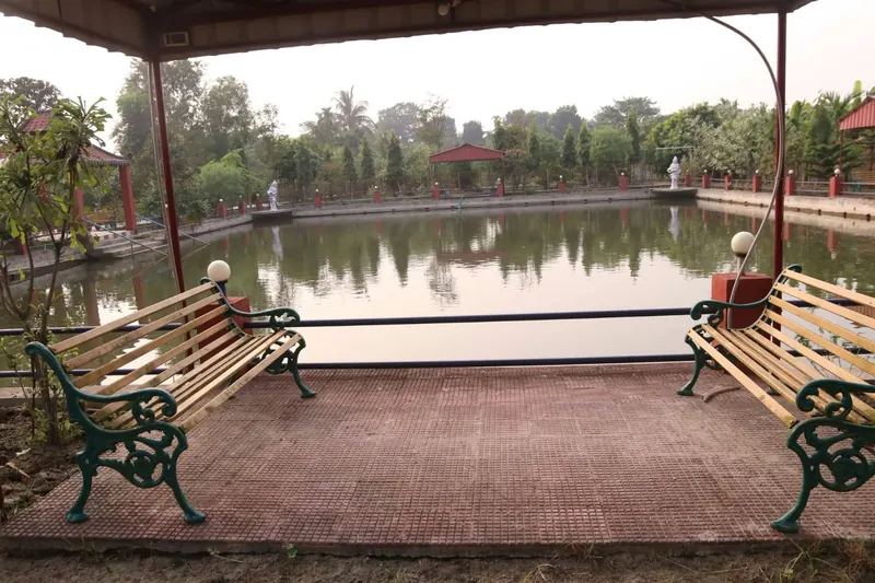 Two park benches under a covered shelter looking out over a still pond lined with trees and red lamp posts