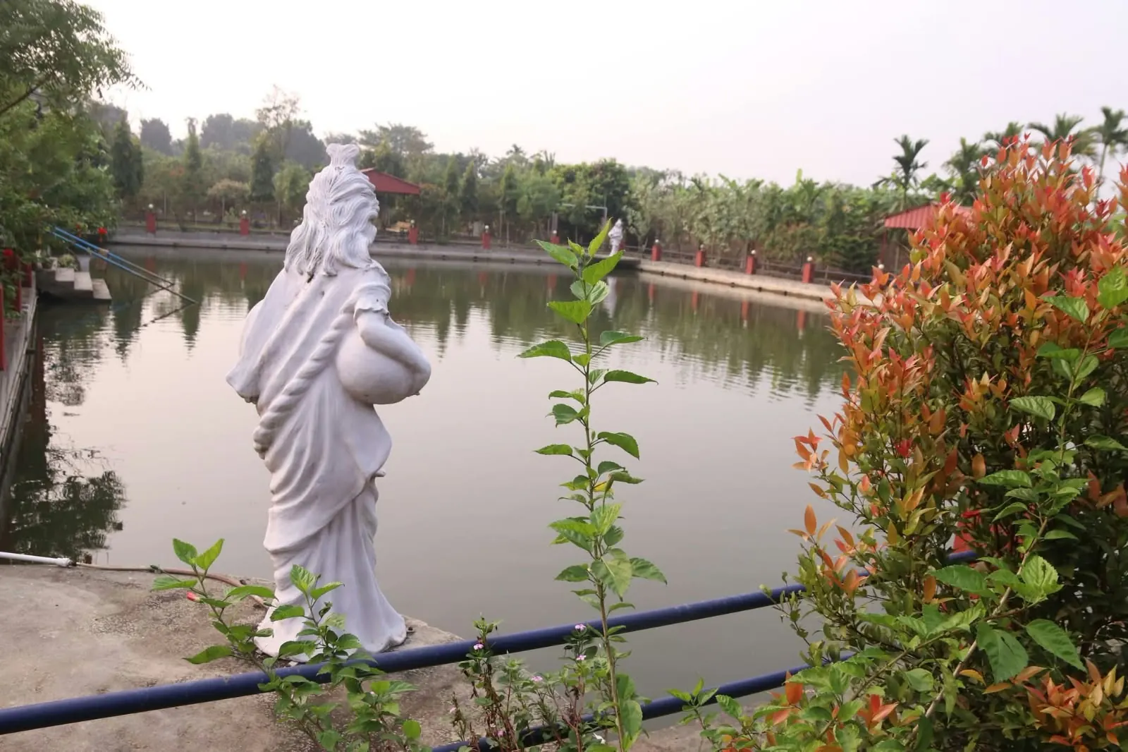 A white statue of a woman carrying a water pot at the edge of a calm pond